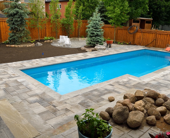 Aménagement Paysager avec Piscine Creusée et Pavé Uni Piscine creusée rectangulaire entourée d'un pavé uni gris dans un jardin avec clôture en bois et arbres.